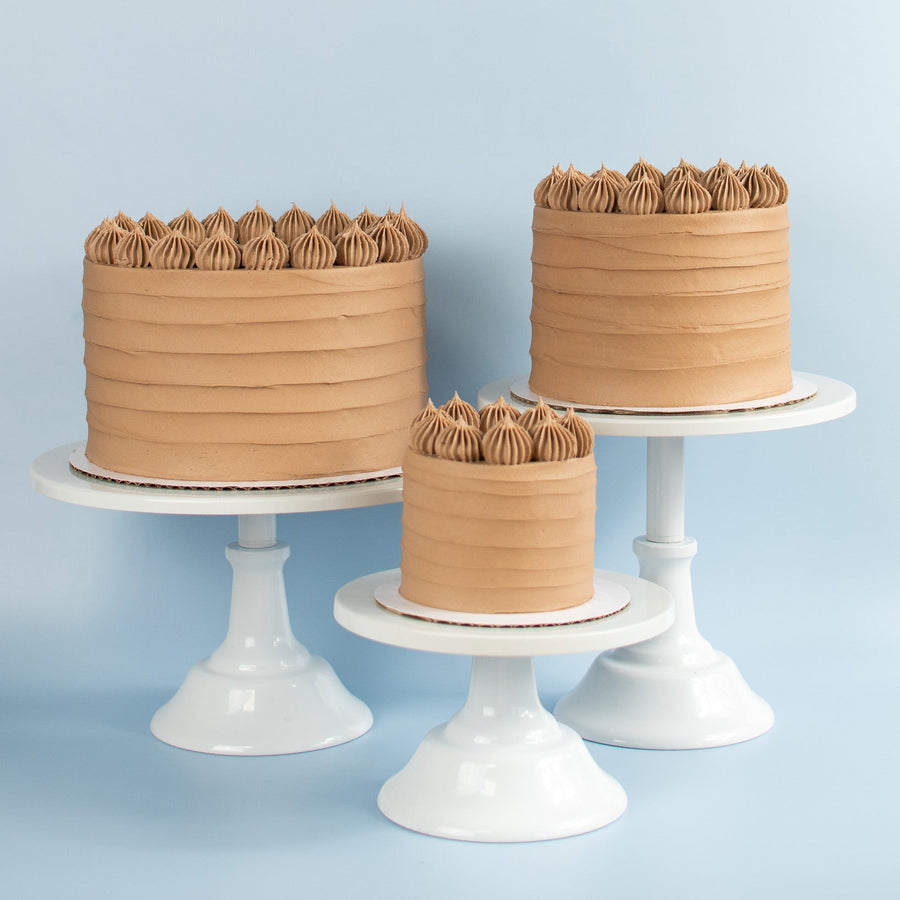 Three chocolate frosted cakes piped decorative frosting tops in different sizes displayed on white pedestal stands against a light blue background.