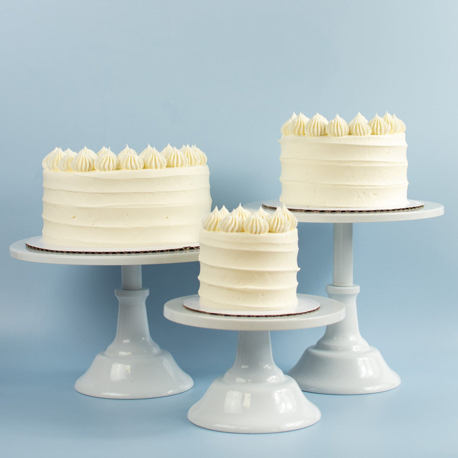 Three vanilla frosted cakes piped decorative frosting tops in different sizes displayed on white pedestal stands against a light blue background.